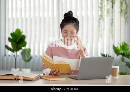 A happy and pretty Asian female college student wearing eyeglasses is reading a book, preparing for the exam, working at her desk in her room. Stock Photo