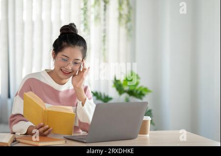 A happy and pretty Asian female college student wearing eyeglasses is reading a book, preparing for the exam, working at her desk in her room. Stock Photo