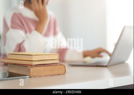 Close-up image of books on a table with a blurred background of a woman working on a laptop. university student, freelancer, remote working Stock Photo