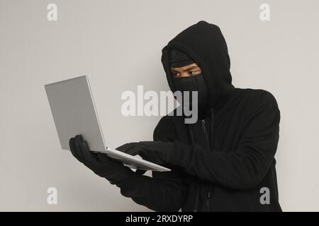 Portrait of mysterious man wearing black hoodie and mask doing hacking activity on laptop, hacker holding a personal computer. Cyber security concept. Stock Photo