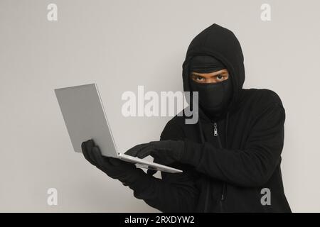 Portrait of mysterious man wearing black hoodie and mask doing hacking activity on laptop, hacker holding a personal computer. Cyber security concept. Stock Photo