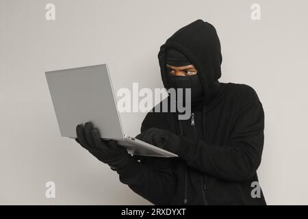 Portrait of mysterious man wearing black hoodie and mask doing hacking activity on laptop, hacker holding a personal computer. Cyber security concept. Stock Photo