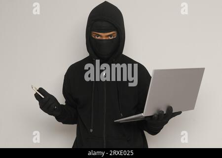 Portrait of mysterious man wearing black hoodie and mask doing hacking activity on laptop, hacker holding a personal computer. Cyber security concept. Stock Photo
