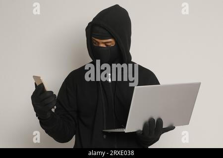 Portrait of mysterious man wearing black hoodie and mask doing hacking activity on laptop, hacker holding a mobile phone. Cyber security concept. Isol Stock Photo