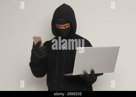 Portrait of mysterious man wearing black hoodie and mask doing hacking activity on laptop, hacker holding a mobile phone. Cyber security concept. Isol Stock Photo