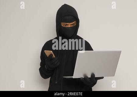 Portrait of mysterious man wearing black hoodie and mask doing hacking activity on laptop, hacker holding a mobile phone. Cyber security concept. Isol Stock Photo
