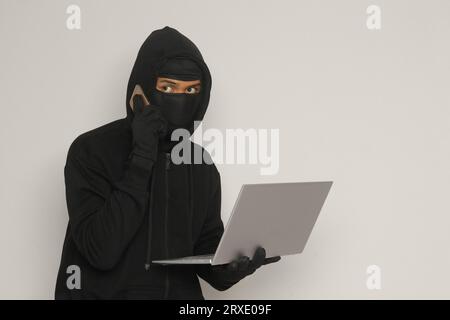 Portrait of mysterious man wearing black hoodie and mask doing hacking activity on laptop, hacker holding a mobile phone. Cyber security concept. Isol Stock Photo