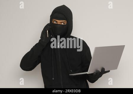Portrait of mysterious man wearing black hoodie and mask doing hacking activity on laptop, hacker holding a mobile phone. Cyber security concept. Isol Stock Photo