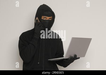 Portrait of mysterious man wearing black hoodie and mask doing hacking activity on laptop, hacker holding a mobile phone. Cyber security concept. Isol Stock Photo