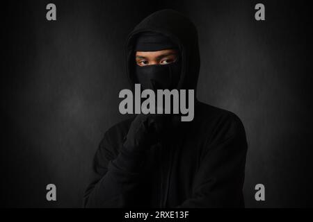 Mysterious man wearing black hoodie and mask standing against dark background, looking at camera. Dramatic low light portrait Stock Photo