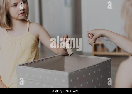 Children cleaning toys from floor after playing, collect them to textile box. Kids sitting on carpet at sunny white home bedroom, kindergarten playroo Stock Photo