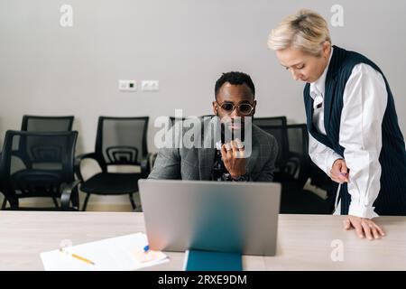 Portrait of female boss mentor helping black male employee with online assignment teach intern in office. Businesswoman executive supervisor helping Stock Photo