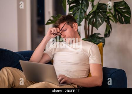 Tired sleepy man rubbing eyes working on laptop sitting on couch in evening at home. Stock Photo