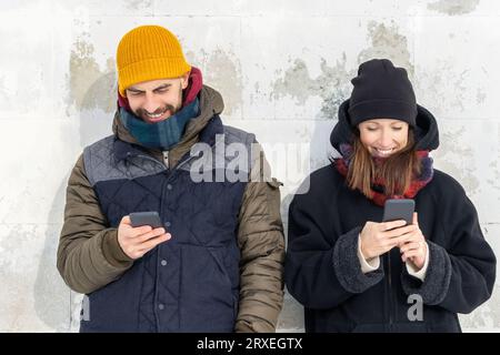 Smiling couple using smartphones on wall background outside. Laughing man and woman standing by white wall holding mobile phones. Positive people man Stock Photo