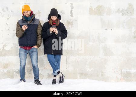 Laughing couple standing by white wall holding mobile phones. Smiling man and woman using smartphones on wall background outside. Positive people man Stock Photo