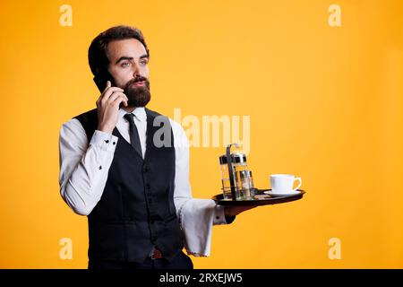 Young waiter talking on phone call with clients to make table reservations for dinner at fancy five star restaurant. Professional stylish butler using mobile phone before serving customers on camera. Stock Photo