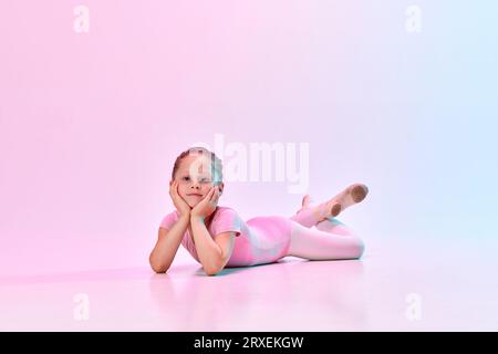 Portrait of small cute ballerina dancer girl wearing ballet swimsuit and pointe lying on floor with head on hands in mixed neon light. Stock Photo