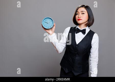 Hotel woman receptionist holding retro alarm clock and looking at camera with serious facial expression. Young asian waitress wearing uniform showing time management concept portrait Stock Photo