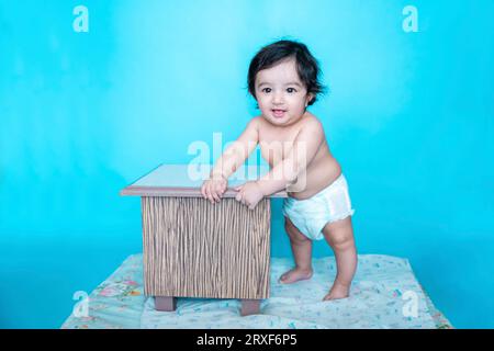 Smiling six months old cute indian baby boy wearing diaper trying to stand with support of wooden stool isolated over blue studio background, Happy ch Stock Photo