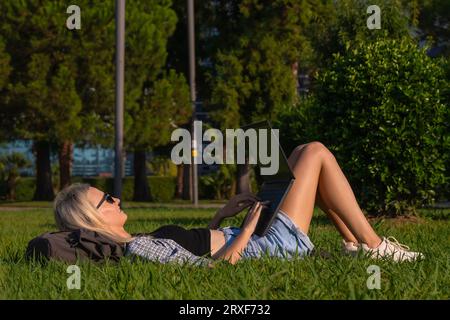 Smiling student uses a laptop lying on the grass. Young woman is lying with a laptop in a park on a sunny day. Stock Photo