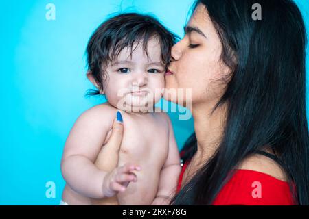 Indian teenager girl kissing six months cute little baby in diaper isolated over blue background. Asian infant child and sister, Happy family. Stock Photo