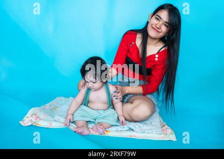 Indian teenager girl sitting with six months cute little baby in diaper isolated over blue background. Asian infant child and sister, Happy family. Stock Photo