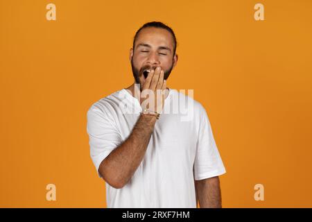 Tired millennial man with beard in white t-shirt covers mouth with hand and yawns Stock Photo
