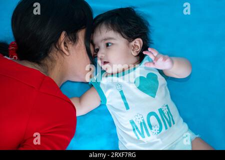 Indian teenager girl lying with six months cute little baby in diaper isolated over blue background. Asian infant child brother and sister, Happy fami Stock Photo