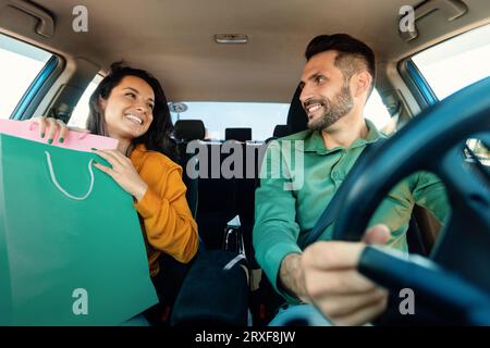 Happy european spouses sitting in car after shopping, woman holding colorful paper shopper bags and smiling Stock Photo
