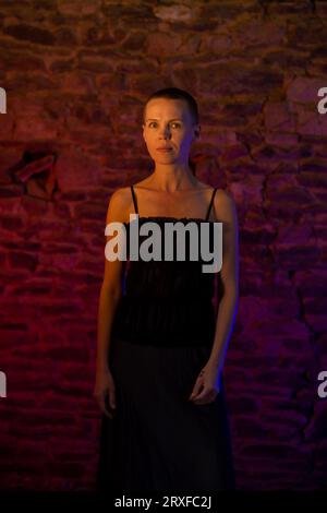 Capturing the poise of a middle-aged lady in her 40s, wearing a black dress, against a brick wall bathed in striking red and blue lighting, her gaze f Stock Photo
