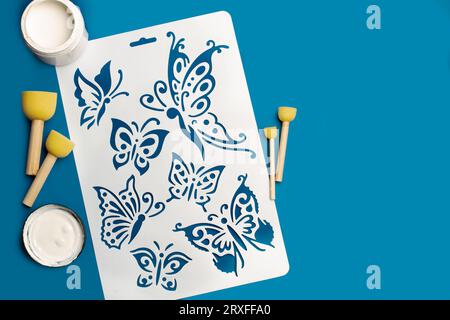 Reusable flexible plastic stencil for graphical design, white paint and round sponge foam brushes on a blue background Stock Photo