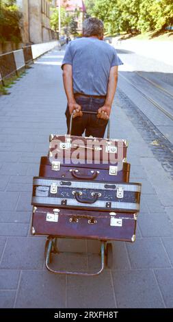 The man pulls old suitcases on a wheelbarrow . Elderly man pulls a wheelbarrow with suitcases. Heap Of Old Suitcases, Trolley Suitcases. The concept o Stock Photo