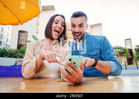Surprised young couple doing online shopping happy to find sales with a cellphone. Mele and female having fun sharing media with a mobile phone. Friends watching a smartphone on a coffee shop terrace. High quality photo Stock Photo