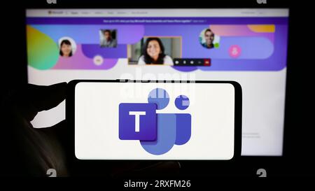 Person holding cellphone with logo of business communication platform Microsoft Teams on screen in front of webpage. Focus on phone display. Stock Photo