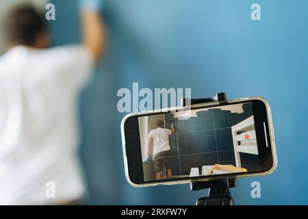 caucasian bearded man painting wall and recording video on smartphone. Painting apartment, renovating home Stock Photo