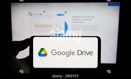 Person holding cellphone with logo of file storage platform Google Drive on screen in front of business webpage. Focus on phone display. Stock Photo