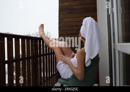 a young beautiful woman after spa treatments, wrapped in a towel, sits on a snow-covered balcony Stock Photo