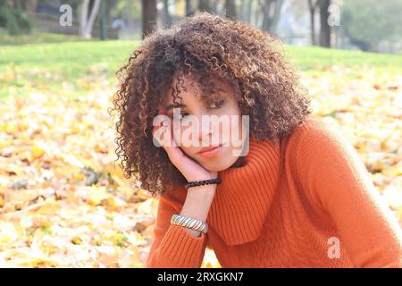 Curly haired woman with orange sweater posing with the autumn leaves in background Stock Photo