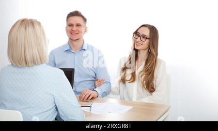 An experienced woman realtor advises a young couple about buying real estate in the office. Selective focus. Stock Photo