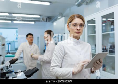 Portrait of a confident young woman doctor scientist standing in a research laboratory, holding a tablet in her hands, looking seriously into the camera. Male colleagues work nearby. Stock Photo