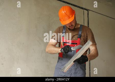Worker in overalls and safety hard hat holds and prepares two spatulas for plastering concrete wall with putty using. Renovation apartment and construction new building concept. Copy place. Stock Photo