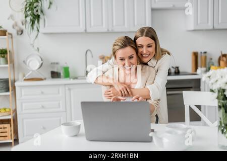 attractive blonde sisters looking at laptop smiling and hugging from behind, family bonding Stock Photo