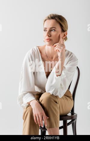 beautiful blonde woman in formal wear sitting on chair and touching her face, style and fashion Stock Photo