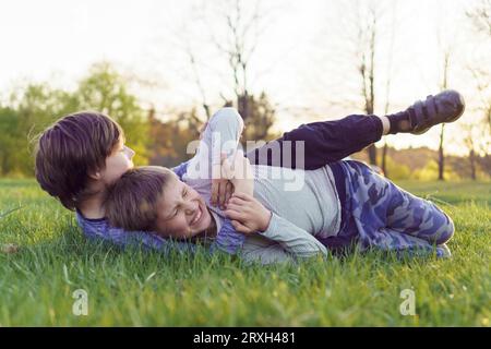 Image capturing funny leisure of two teenagers lying on green meadow, fighting for fun and playing. Boy in blue longsleeve trying to grab and Stock Photo