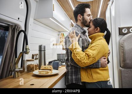 Side view portrait of young couple embracing affectionately with husband giving kiss on forehead while standing in trailer van and travelling together, copy space Stock Photo