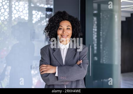 Portrait of young African American businesswoman, woman with crossed arms smiling and looking at camera, female worker at workplace inside office, boss in business suit smiling with satisfaction. Stock Photo