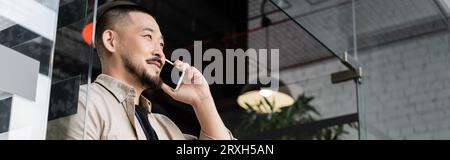 happy asian man having business phone call on smartphone near glass door in office, banner Stock Photo