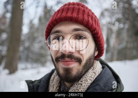 Close up portrait of bearded young man wearing glasses looking at camera outdoors in winter forest Stock Photo
