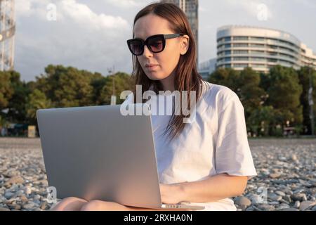 Digital nomad woman in sunglasses working with laptop sits on the beach by the sea during sunset. Male freelancer work online from a distance. Remote Stock Photo