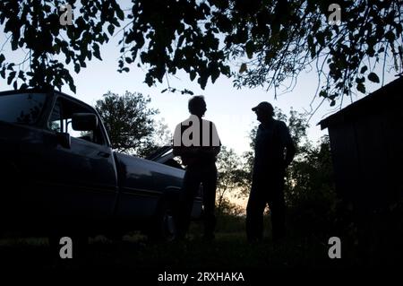 Two farmers are silhouetted against the setting sun as they stand together and talk beside a barn and pickup truck Stock Photo